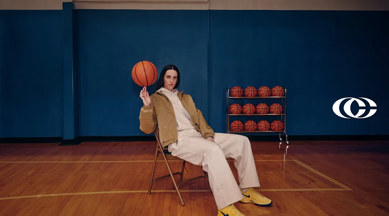 Nike Caitlin 1 Collection campaign image featuring an athlete in a khaki jacket, sail sweatsuit and yellow basketball shoes seated on an indoor basketball court.